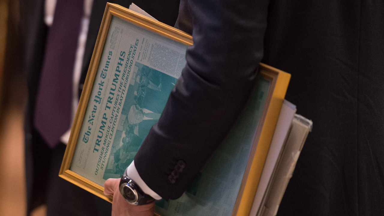 An aide to then-president-elect Donald Trump carries a framed New York Times front page featuring his election win. Critics are attacking the NYT for creating an interactive article featuring insults he has directed at people over the years.