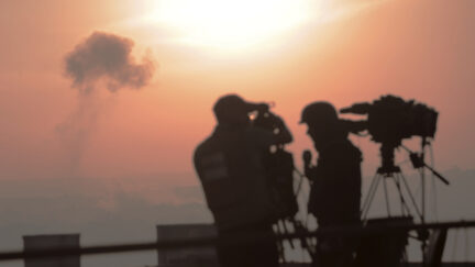 Palestinian journalists are working with an image of an Israeli airstrike in the background.